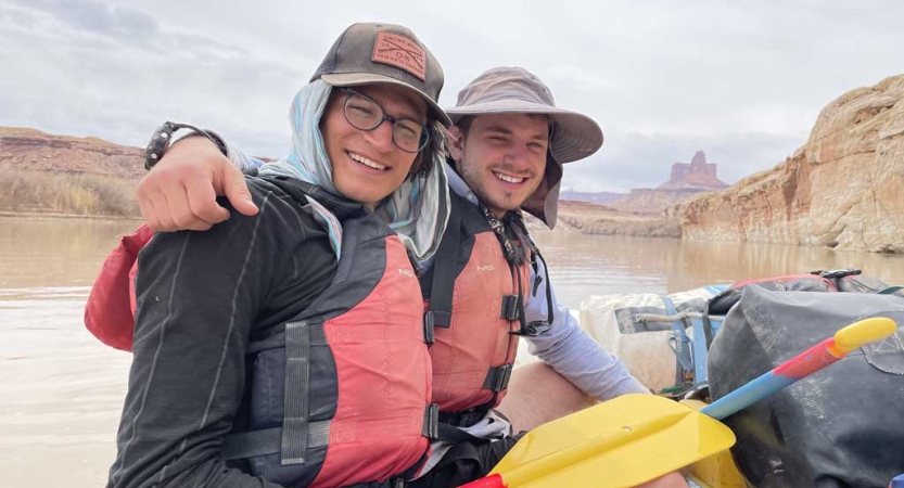 Two people wearing life jackets and hats smile while sitting on a raft. Behind them is the calm river and tall canyon walls. 